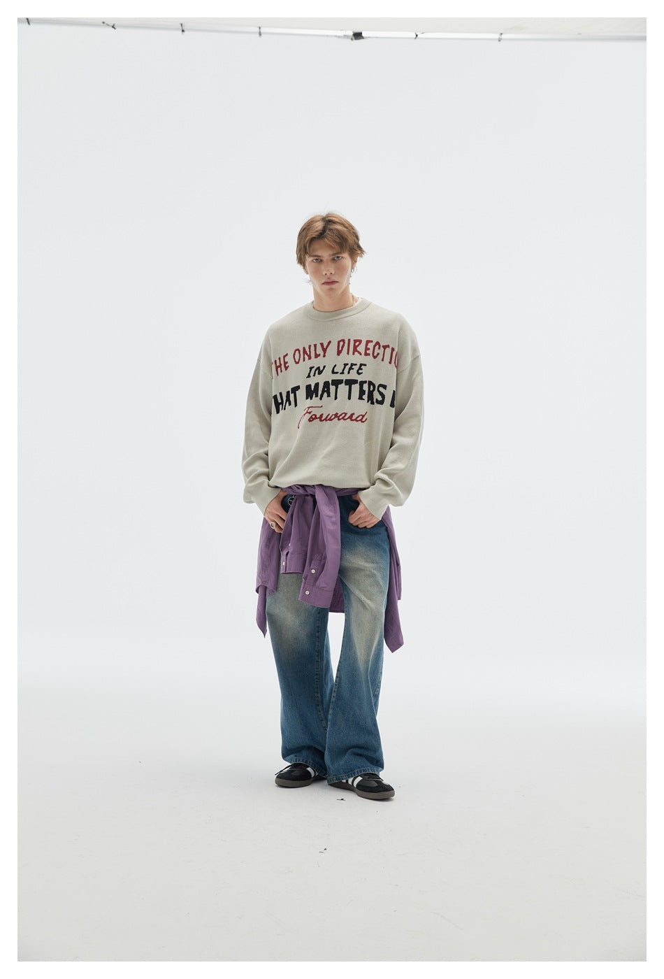 Front view of a model wearing a light gray slogan sweatshirt, blue jeans, and a purple shirt tied at the waist.