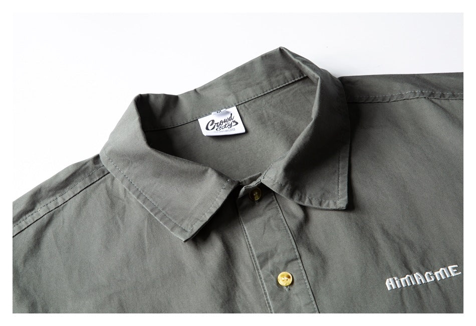 Close-up of a gray-green shirt collar, gold-toned buttons, sewn neck label, and white chest embroidery.