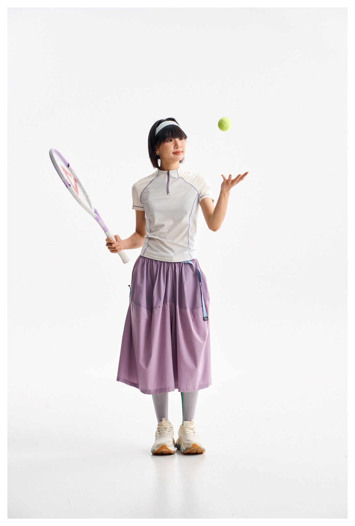 Model holding a tennis racket in a white zip-neck top with purple piping and a lavender skirt.