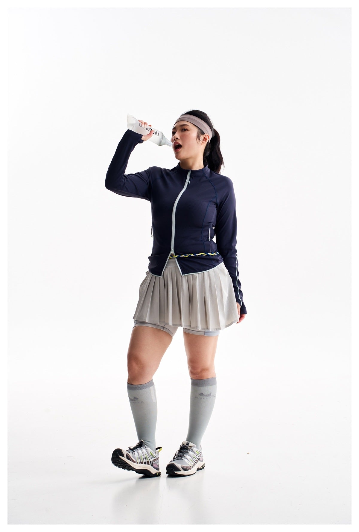 Model drinking from a bottle while wearing a navy zip-up jacket, gray pleated skort, and gray knee-high socks.