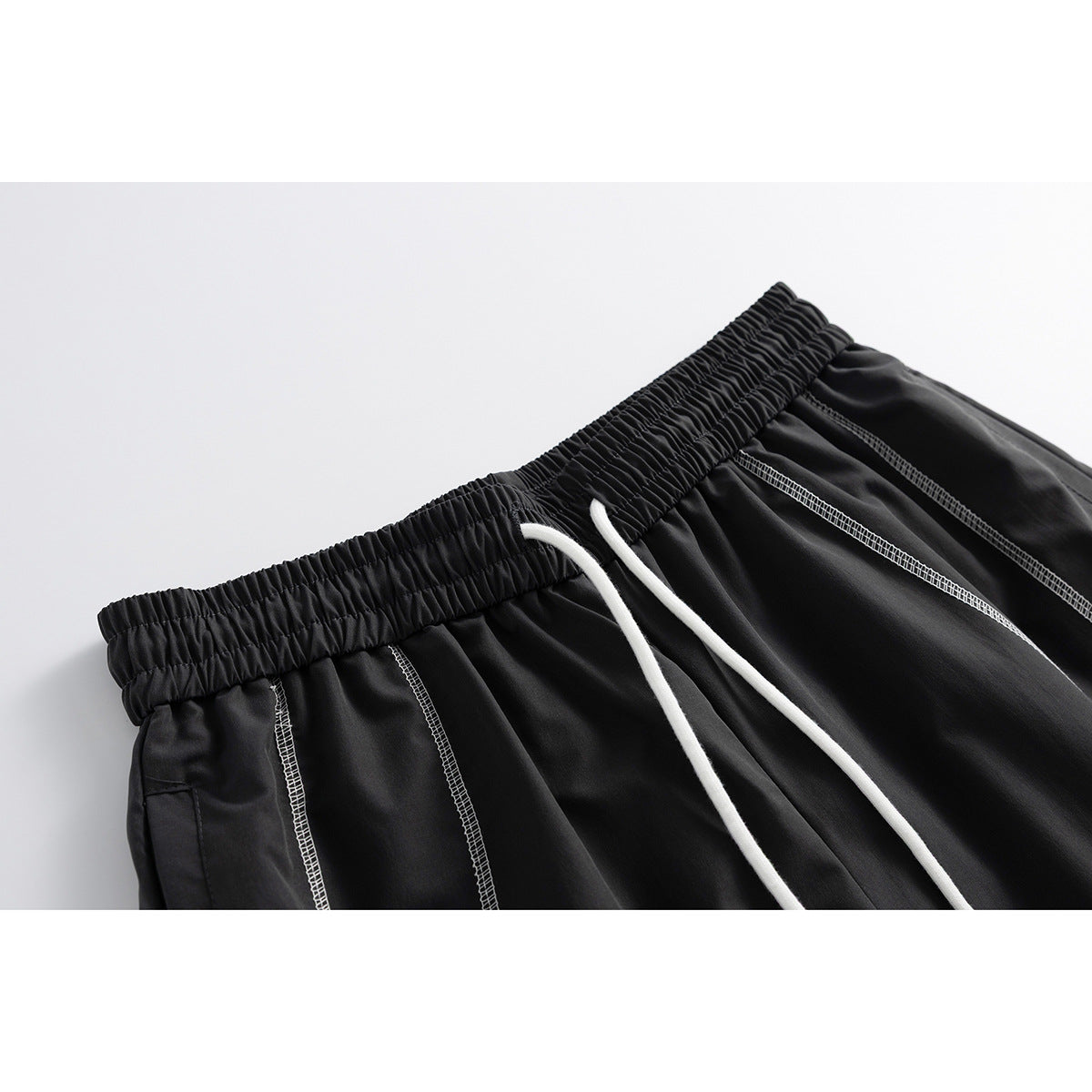 Close-up of black shorts with a gathered elastic waistband, white drawstrings, and contrasting white seam stitching.