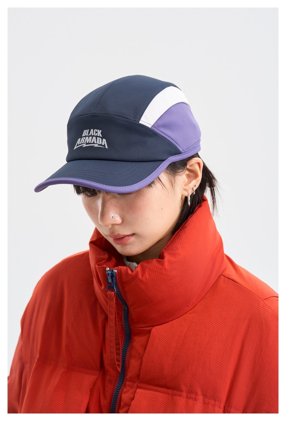 Close-up of a model wearing a red-orange puffer jacket and a navy, white, and purple cap with Black Armada lettering.
