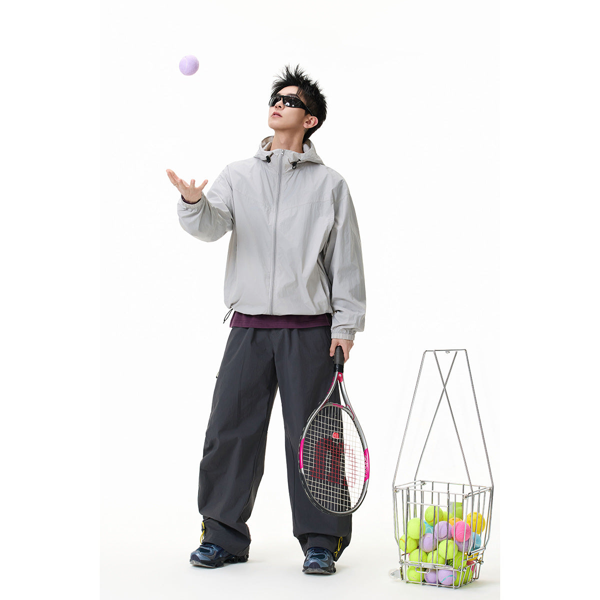 Model wearing a light gray hooded zip jacket and dark wide-leg pants, holding a tennis racket beside a basket of balls.
