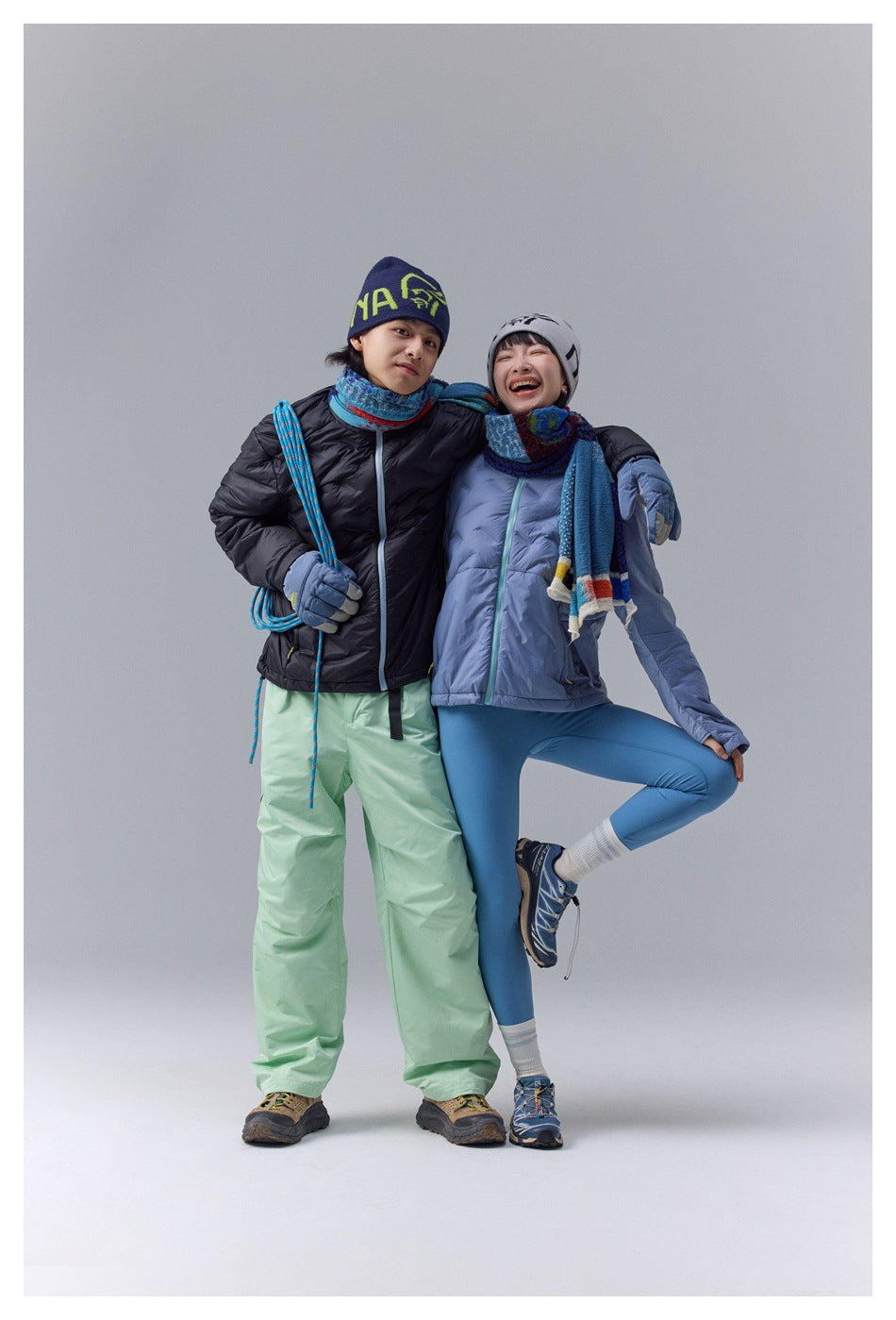 Two models pose together wearing black and light blue quilted zip-front jackets with scarves and winter accessories.