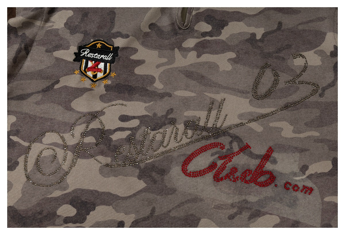 Close-up of gray-brown camouflage fabric with a stitched crest and decorative rhinestone lettering.
