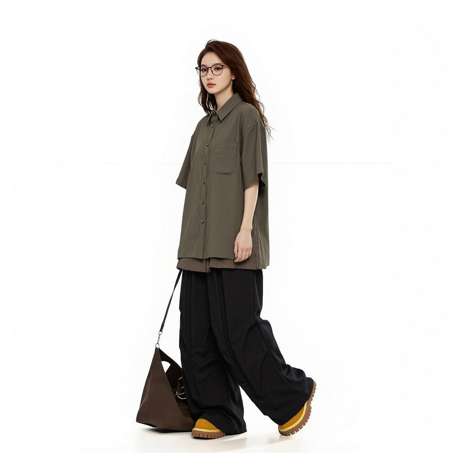 Woman in an olive short-sleeve button-up shirt with a chest pocket and loose black trousers, shown at an angle.