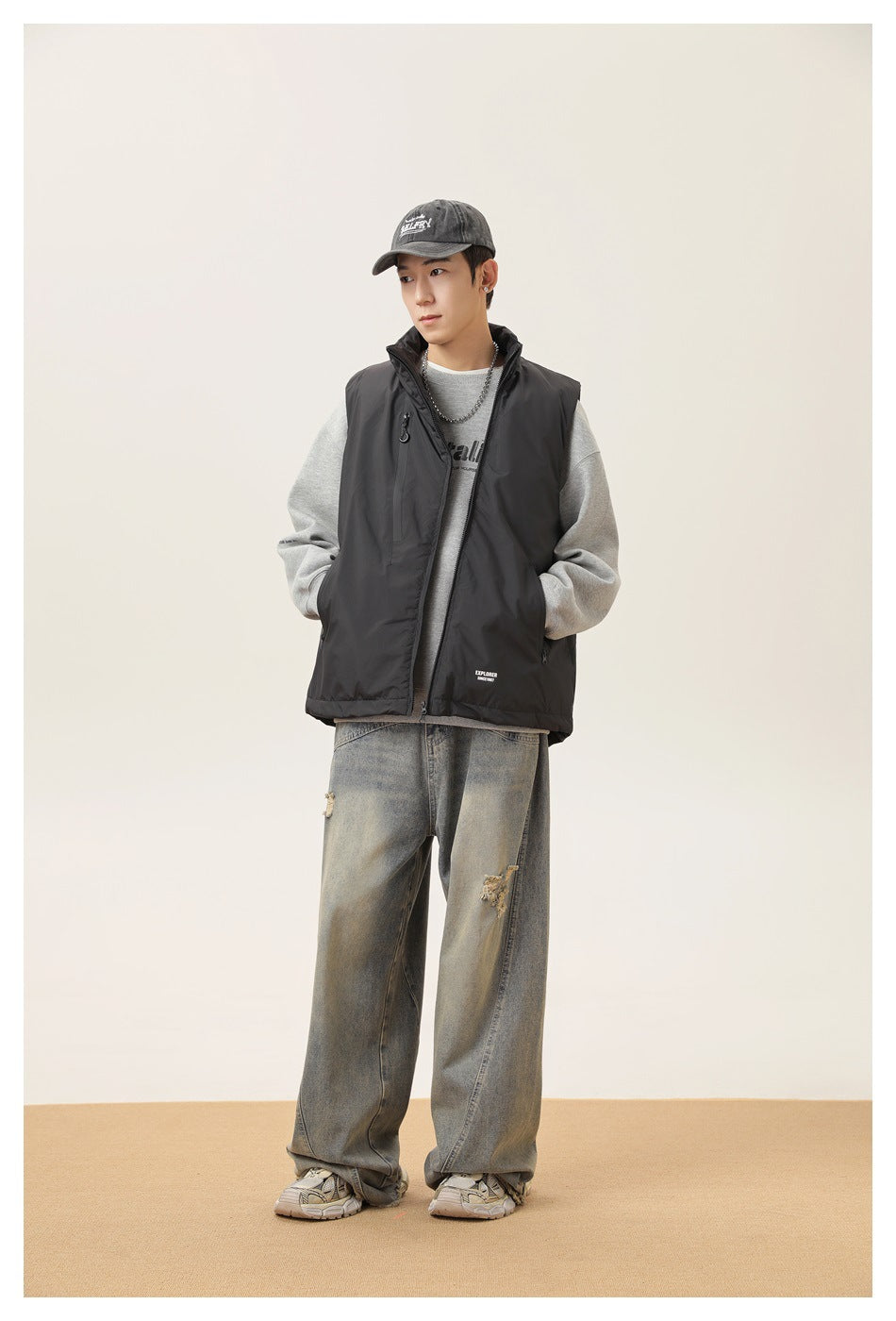 Model wearing a black stand-collar zip vest over a gray sweatshirt with wide-leg faded jeans and a cap