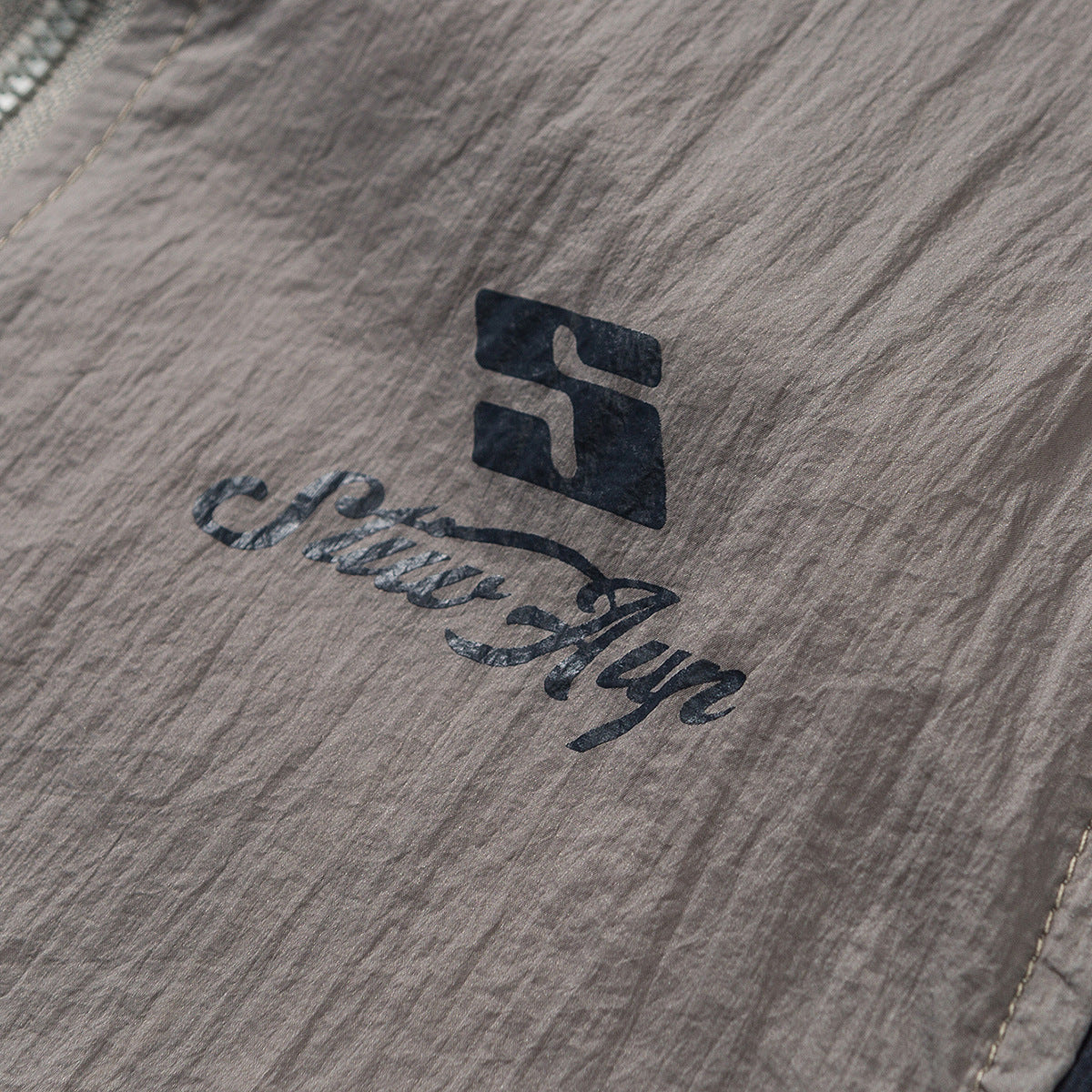 Close-up of a dark printed logo on crinkled taupe-gray garment fabric.