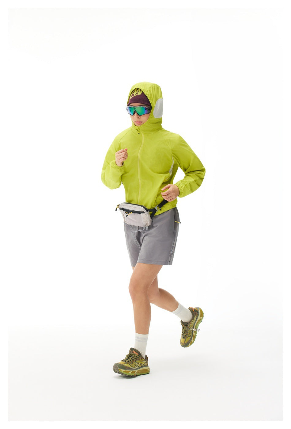 Model jogging in a bright lime hooded zip jacket, gray shorts, and a light waist pack.