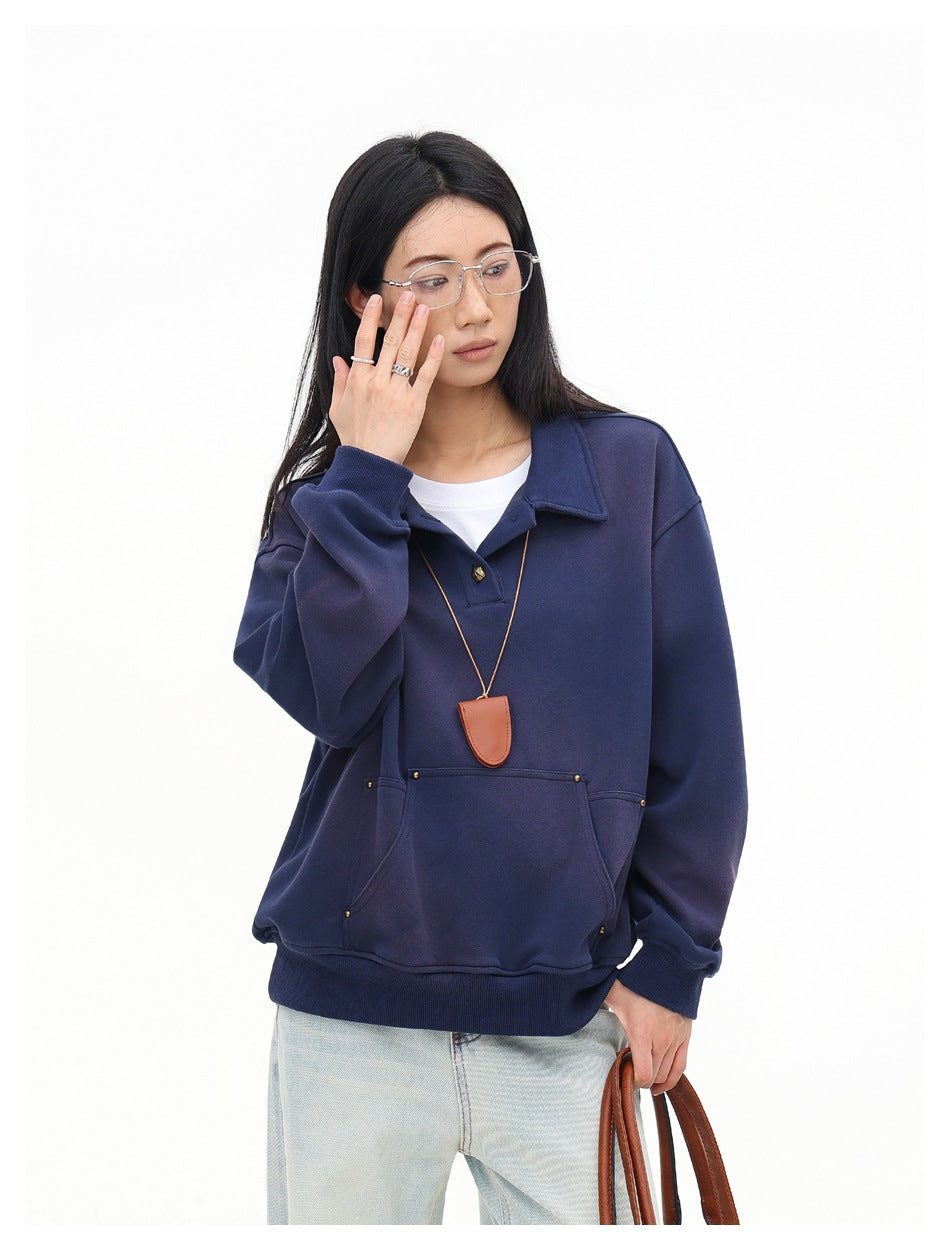 Model wearing an oversized navy collared pullover with a pouch pocket, shown with one hand near her glasses.