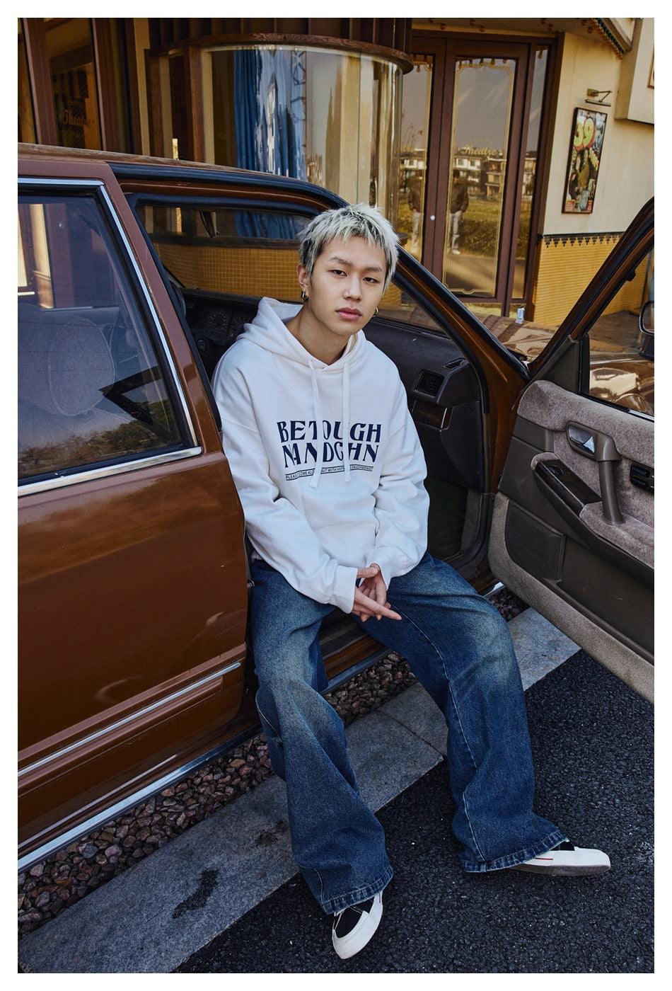 Model seated by an open car door wearing a white graphic pullover hoodie and loose blue jeans.
