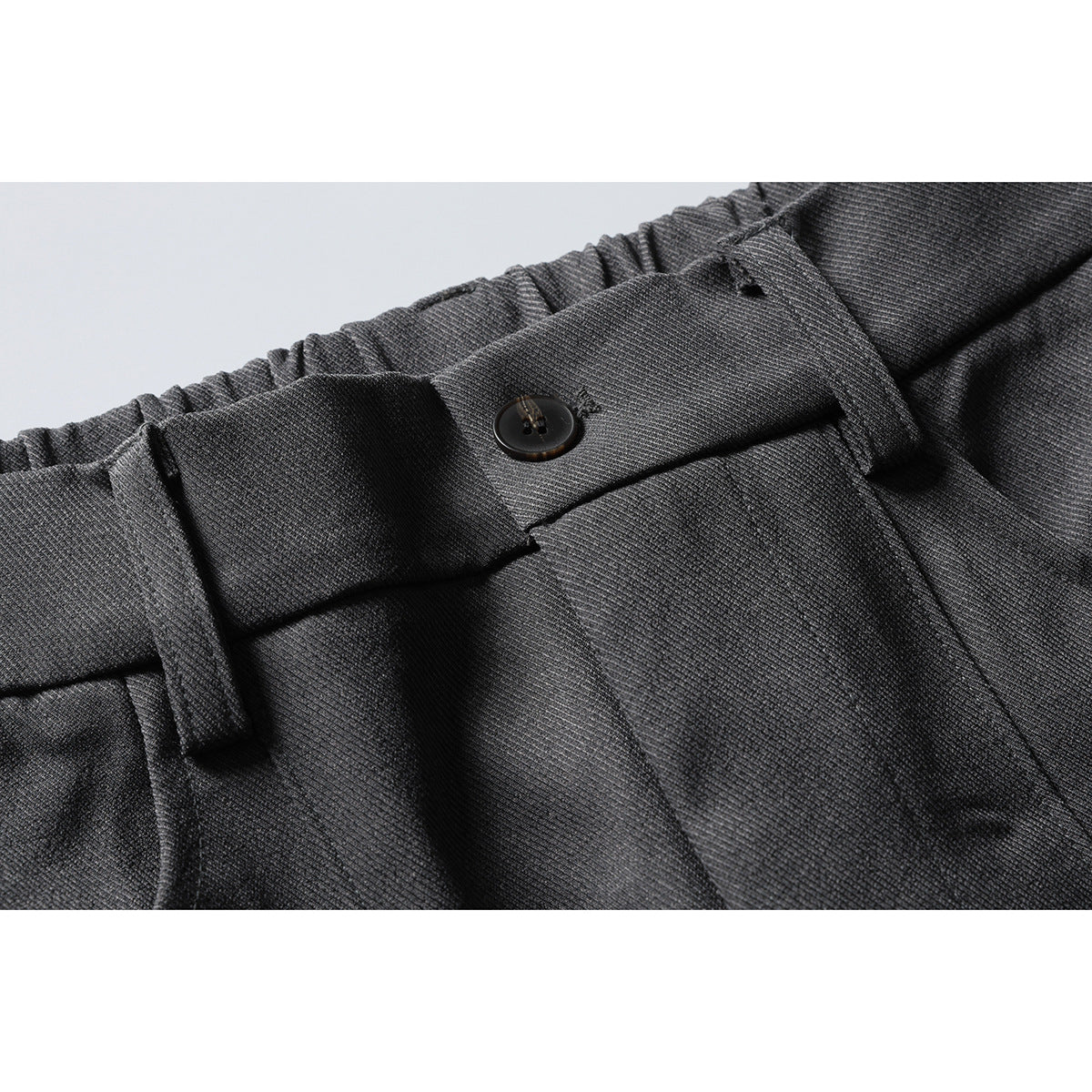 Close-up of charcoal trousers showing an elastic waistband, belt loop, button closure, and front pleats.