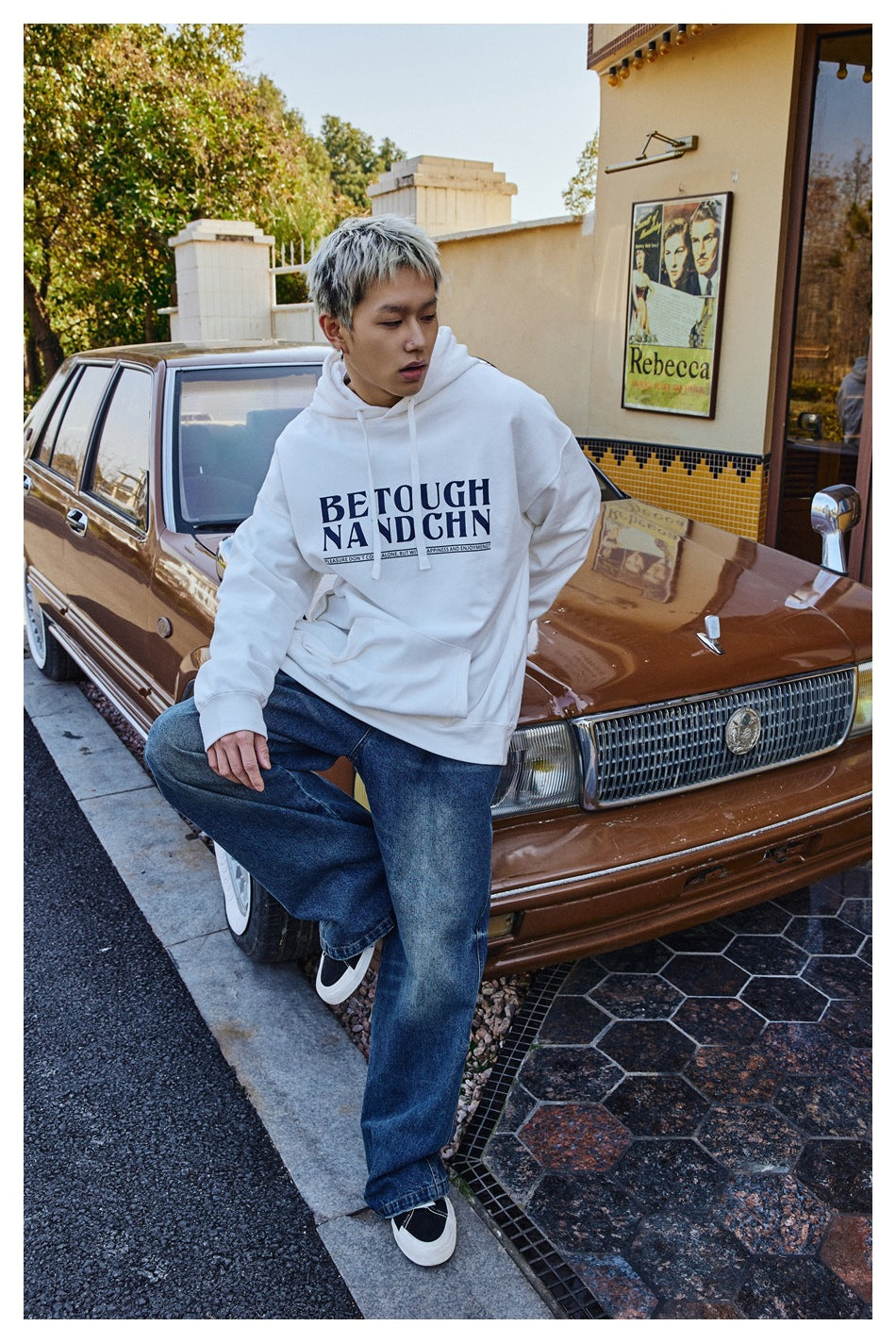 Person wearing a white hoodie with navy chest print and wide-leg jeans, leaning on a vintage brown car