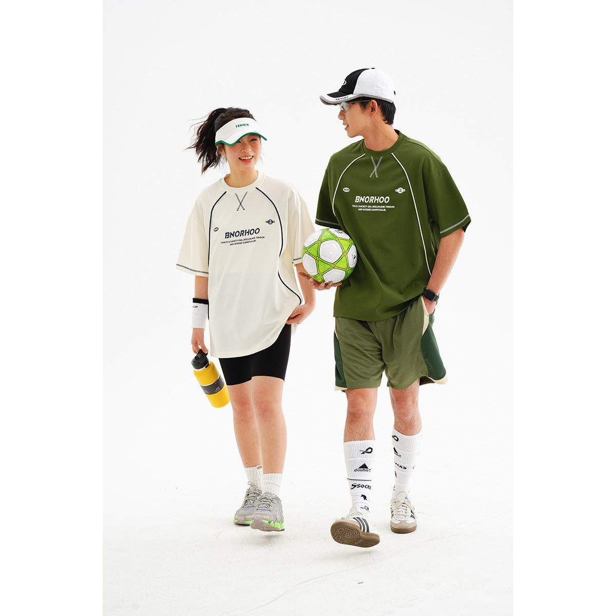 Two models wearing oversized white and olive green athletic T-shirts with matching shorts; one holds a soccer ball.