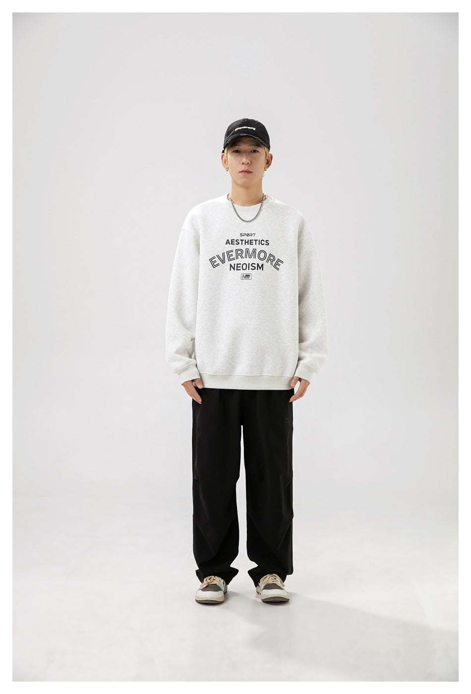 Man wearing light gray crewneck sweatshirt with front lettering print, black pants, cap and sneakers, front view