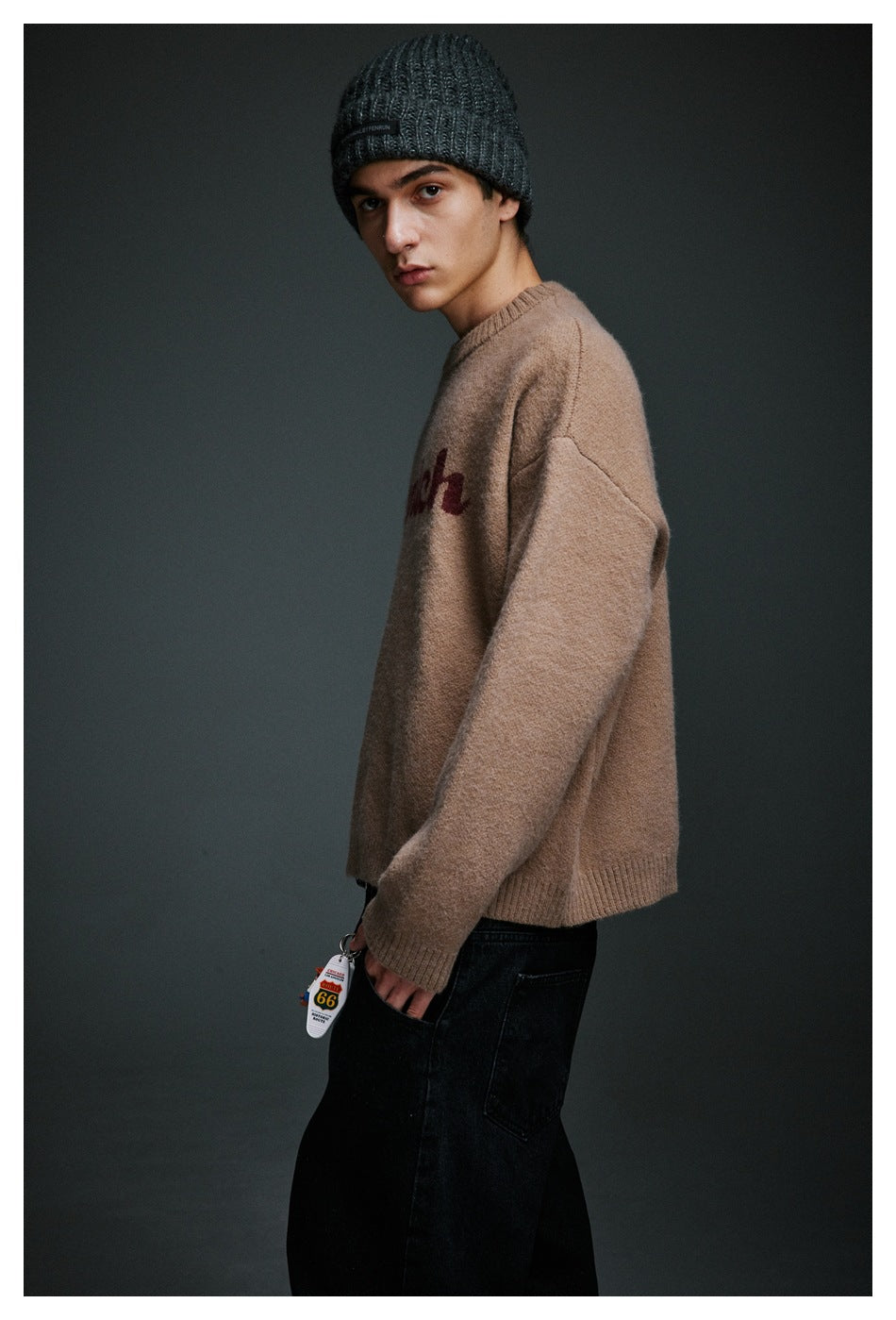 Side view of a model wearing a taupe lettering sweater, black jeans, and a dark gray ribbed beanie.