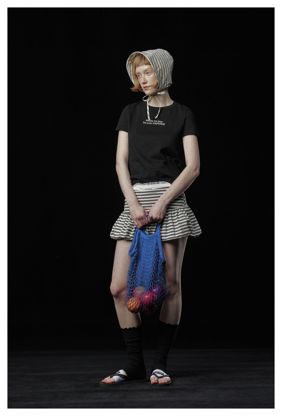 Model in a black short-sleeve slogan T-shirt with striped skirt, bonnet and blue mesh bag, looking sideways