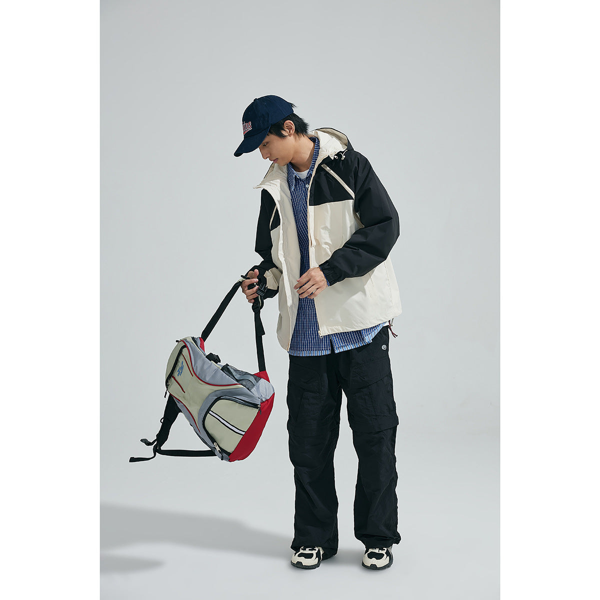 Model wearing a black-and-cream hooded jacket with a blue checked shirt and black cargo pants, holding a bag.