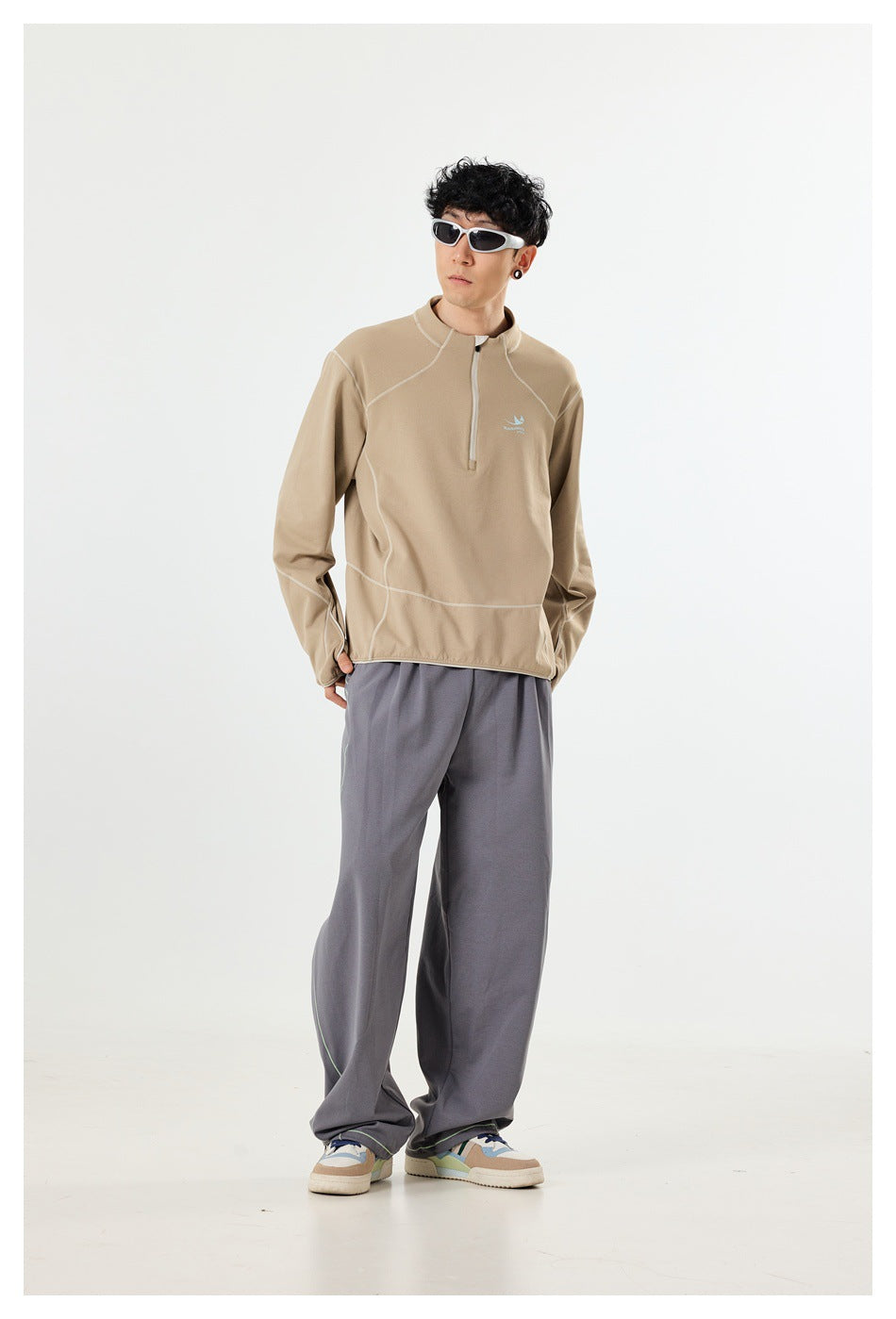 Full-length view of a man wearing a beige quarter-zip pullover with pale contrast seams and loose gray pants.