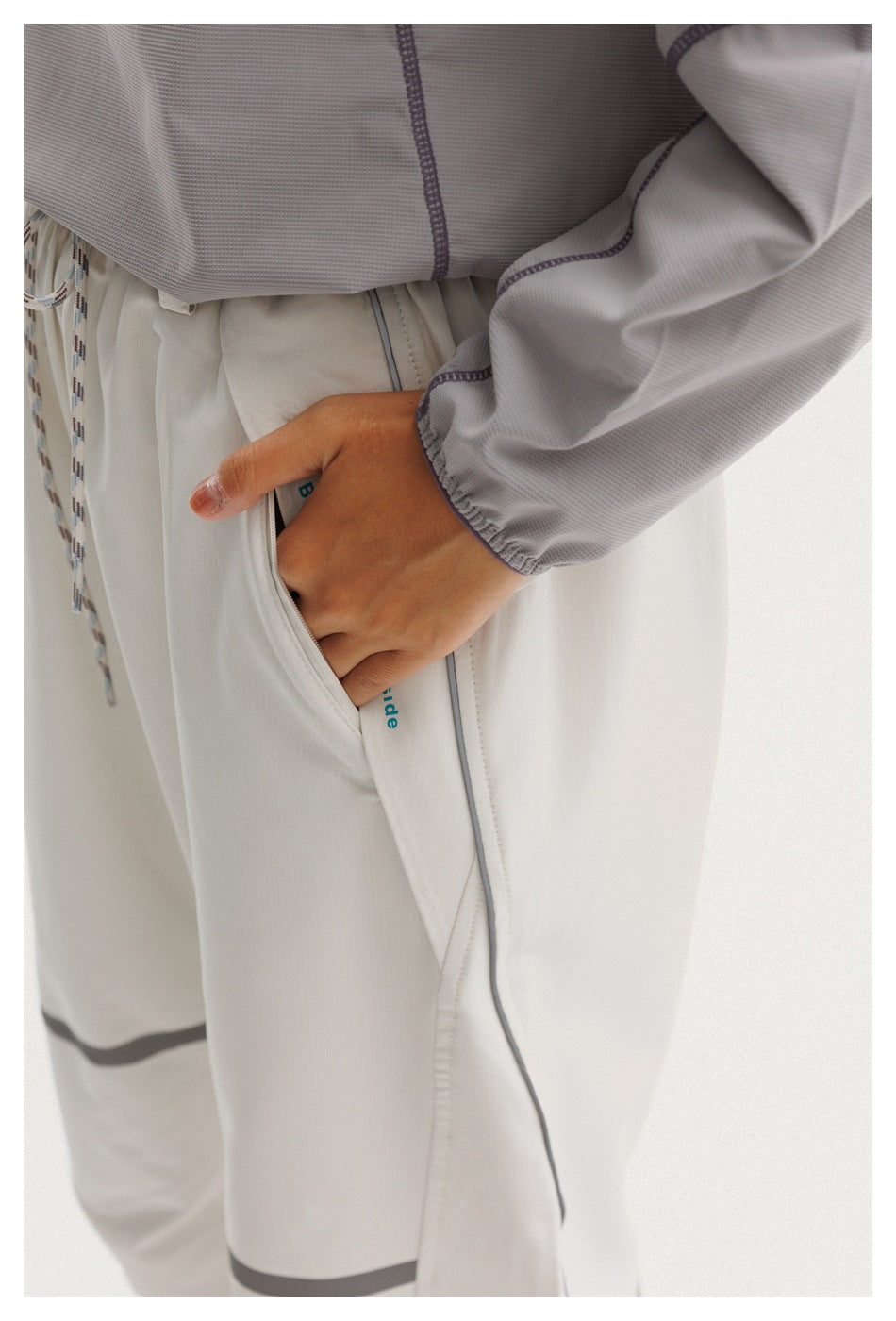 Close-up of light-gray pants showing the pocket, drawstring, piping, and a hand tucked inside.