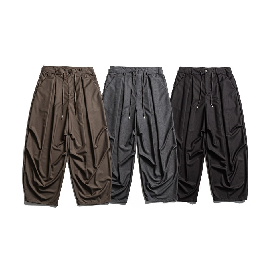 Three pairs of relaxed trousers shown from the front in brown, gray, and black, with pleats and drawstrings.