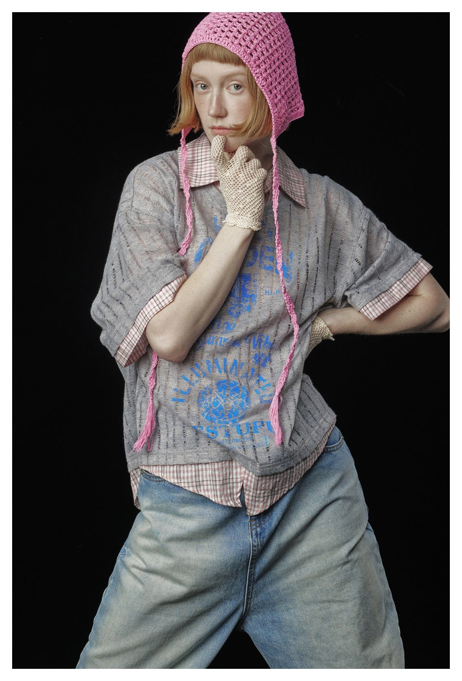 Close view of a model wearing a gray blue-print T-shirt layered over a pink checked shirt, with a pink knit bonnet.