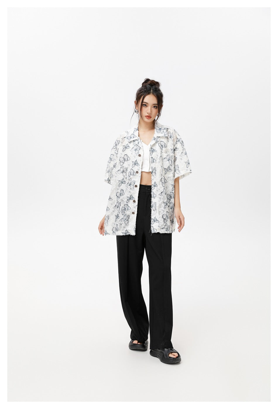 Model wearing an open, oversized white short-sleeve shirt with gray butterfly print over a white crop top and black wide-leg