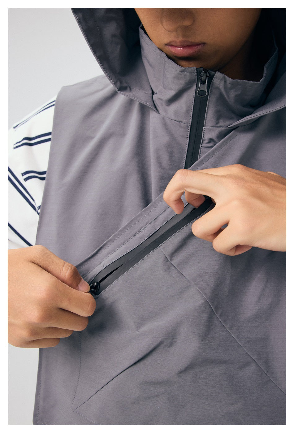 Close-up of the gray vest's hood and front zipper, with hands opening its diagonal zippered chest pocket.
