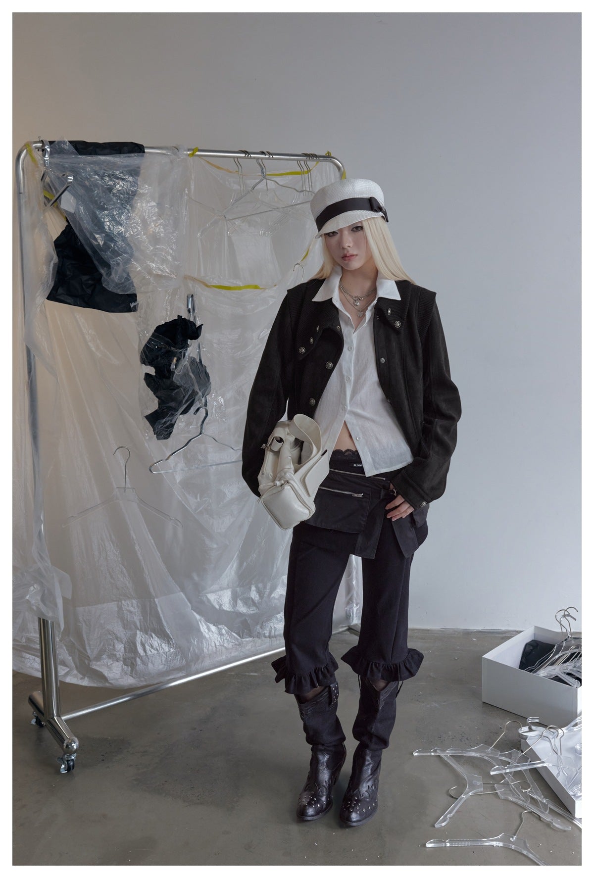 Model wearing the black cropped jacket over an open white shirt with a white cap, black pants and boots, holding a white bag