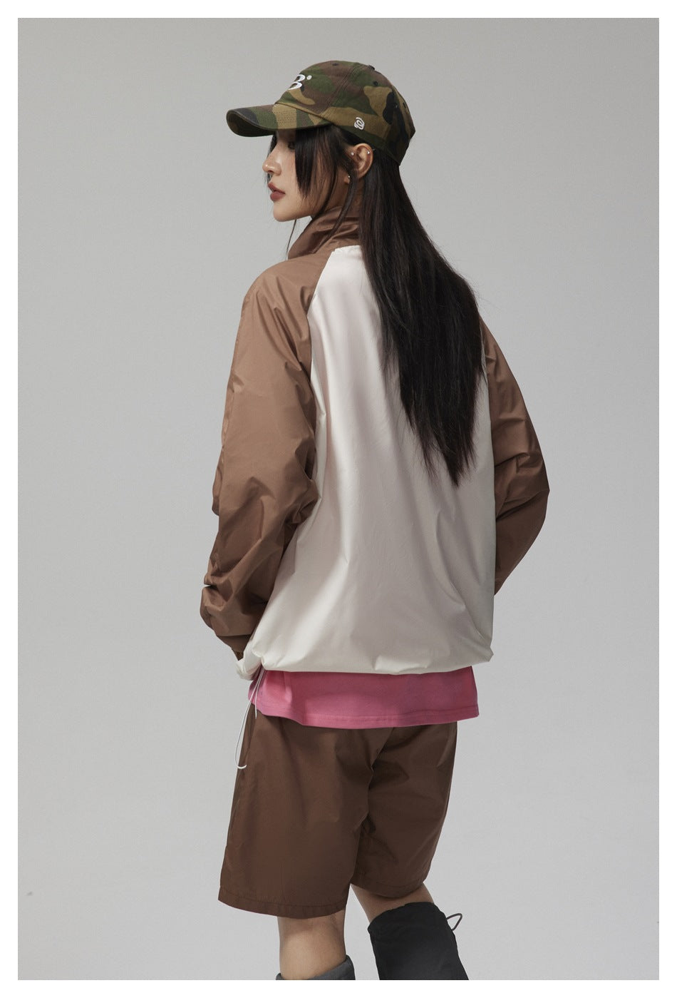 Rear three-quarter view of a person in a white jacket with brown sleeves, brown shorts, and a camouflage cap.
