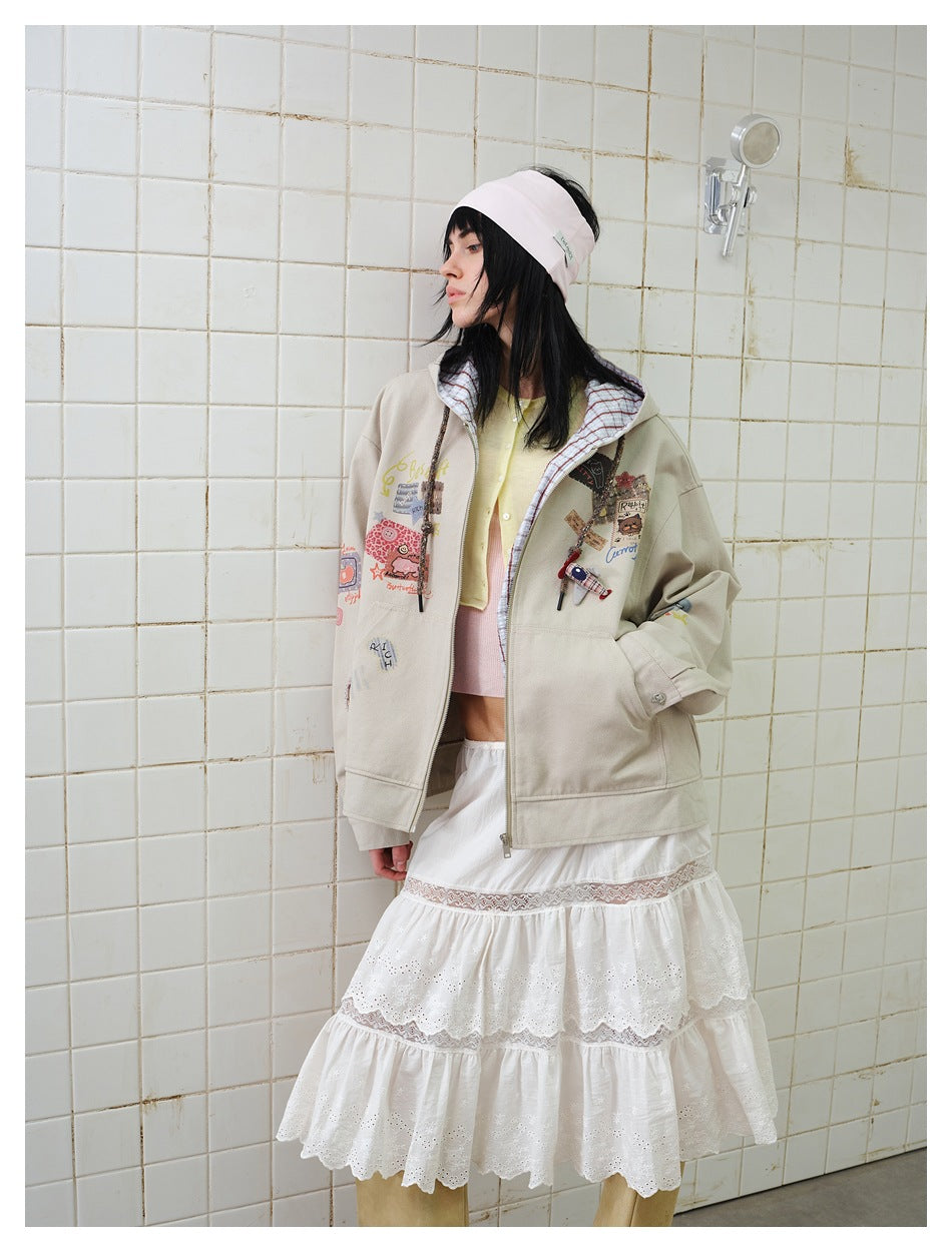 Side-facing model wearing a taupe graphic zip hoodie with plaid hood lining and a white tiered lace skirt.