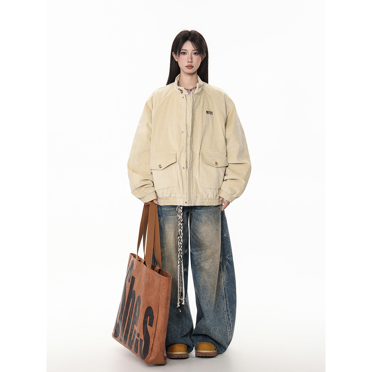 Model in a beige oversized jacket with flap pockets and small chest embroidery, baggy blue jeans, and carrying a brown tote.