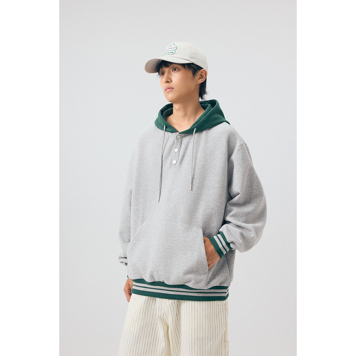 Model wearing a light gray pullover hoodie with a dark green hood, striped cuffs and hem, and a front pouch pocket.