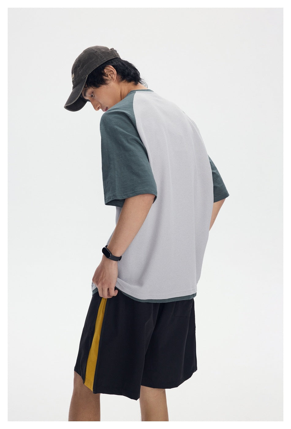 Back view of a model wearing a gray raglan shirt with dark green sleeves and black shorts with a yellow side stripe.