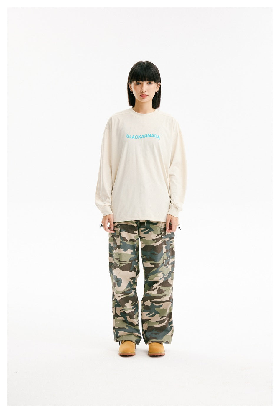 Model wearing a cream long-sleeve shirt with cyan chest lettering and loose camouflage cargo pants.