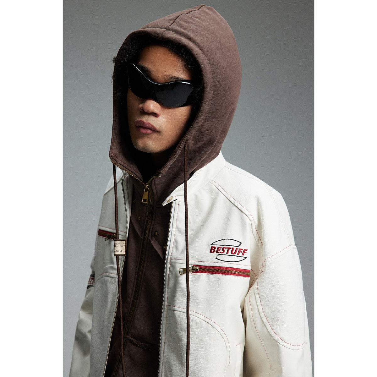 Model wearing a white jacket with red details over a brown hooded layer and black sunglasses.