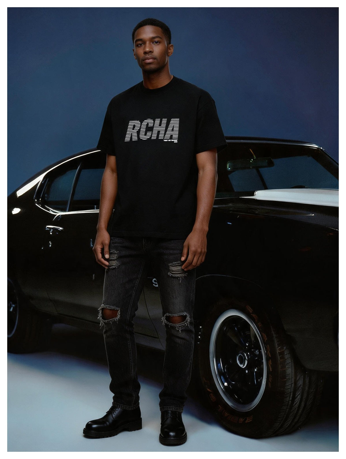Man wearing a black RCHA graphic T-shirt with ripped black jeans and black boots, standing in front of a black car