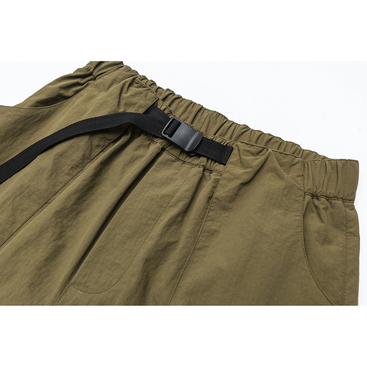 Close-up of olive pants with an elastic waistband, black webbing belt and buckle, and curved pocket seams.