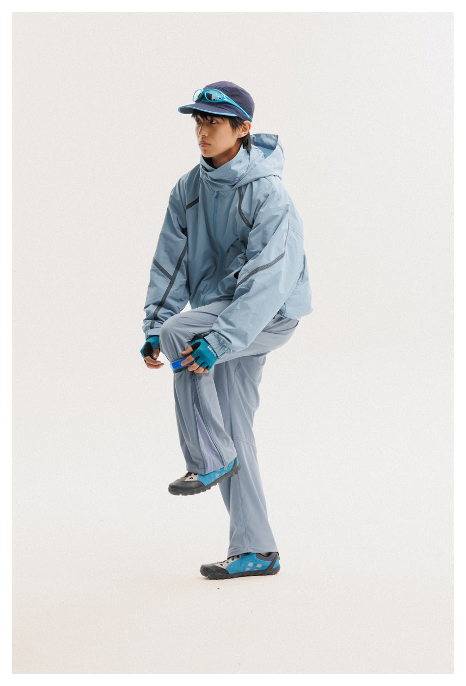 Man in a blue-gray hooded jacket and pants, lifting one leg to adjust the lower-leg cuff.