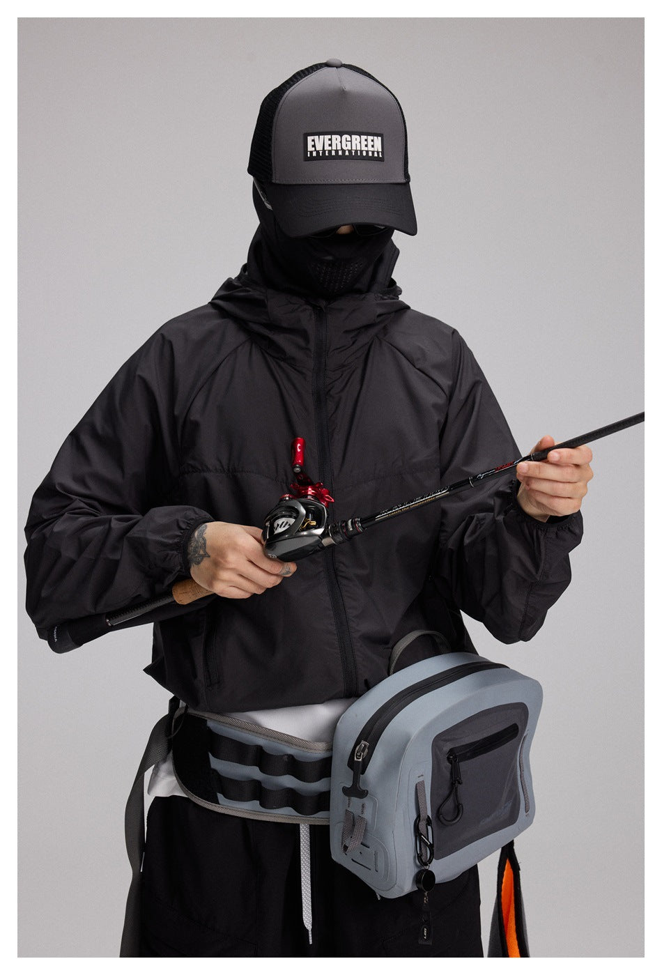 A masked person in a black hooded jacket holds a fishing rod, with a gray fishing bag strapped at the waist.