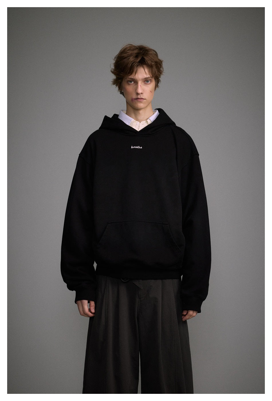 Model wearing a black oversized pullover hoodie over a collared shirt with dark wide-leg trousers against a gray backdrop