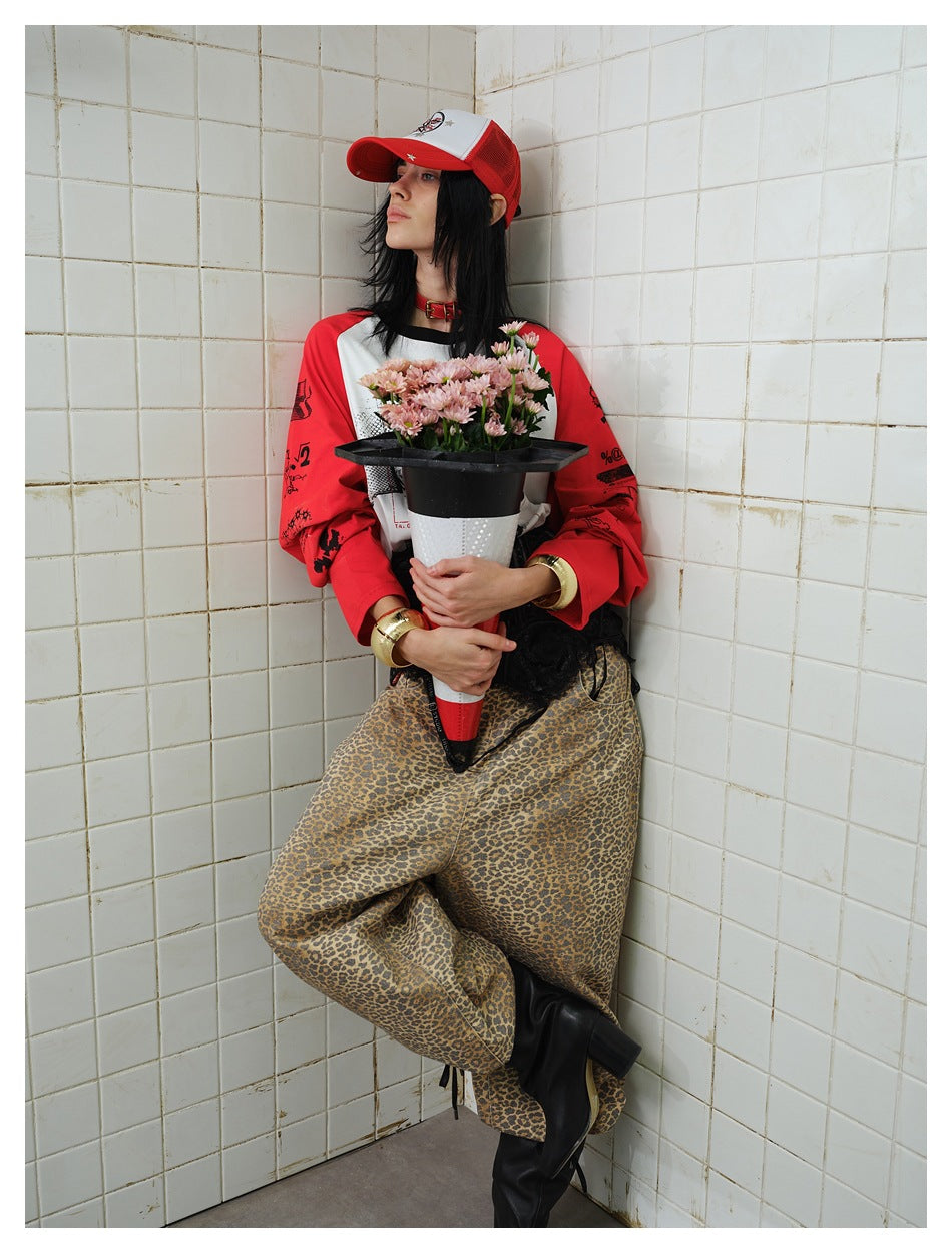 Model leaning in a tiled corner in a red and white raglan top with sleeve prints, cap, leopard pants and black boots