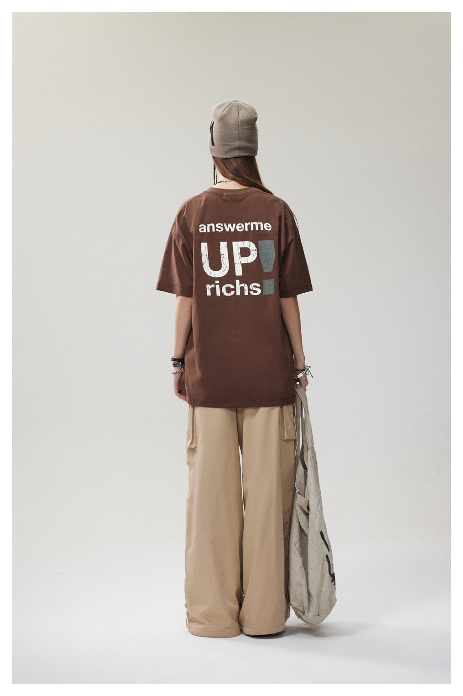 Back view of a model in a brown oversized T-shirt with large white back lettering, beanie and beige cargo pants