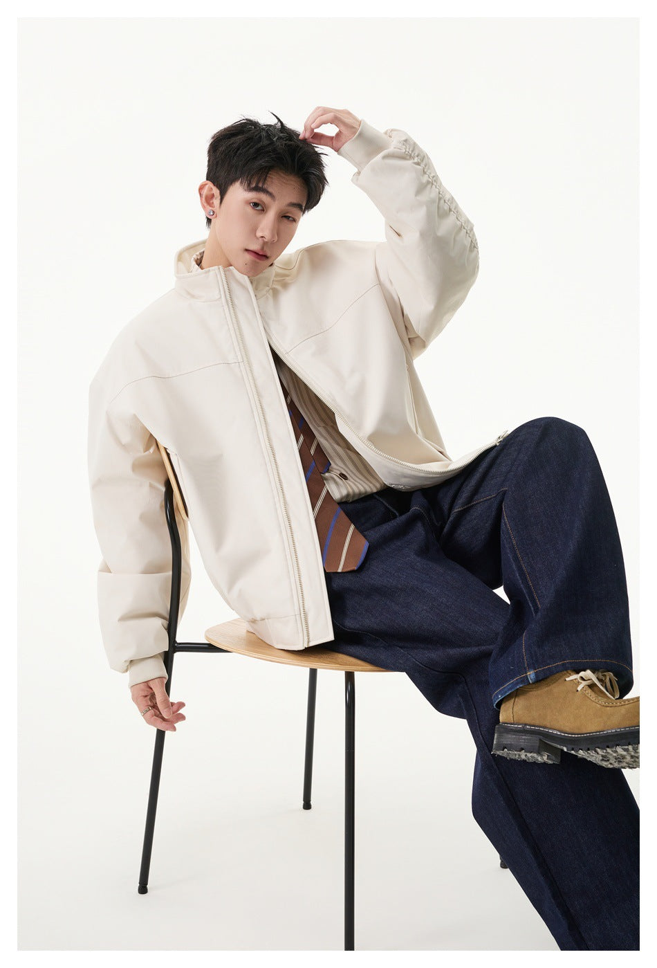 Seated model wearing an open cream jacket over a striped shirt and tie, with dark jeans.