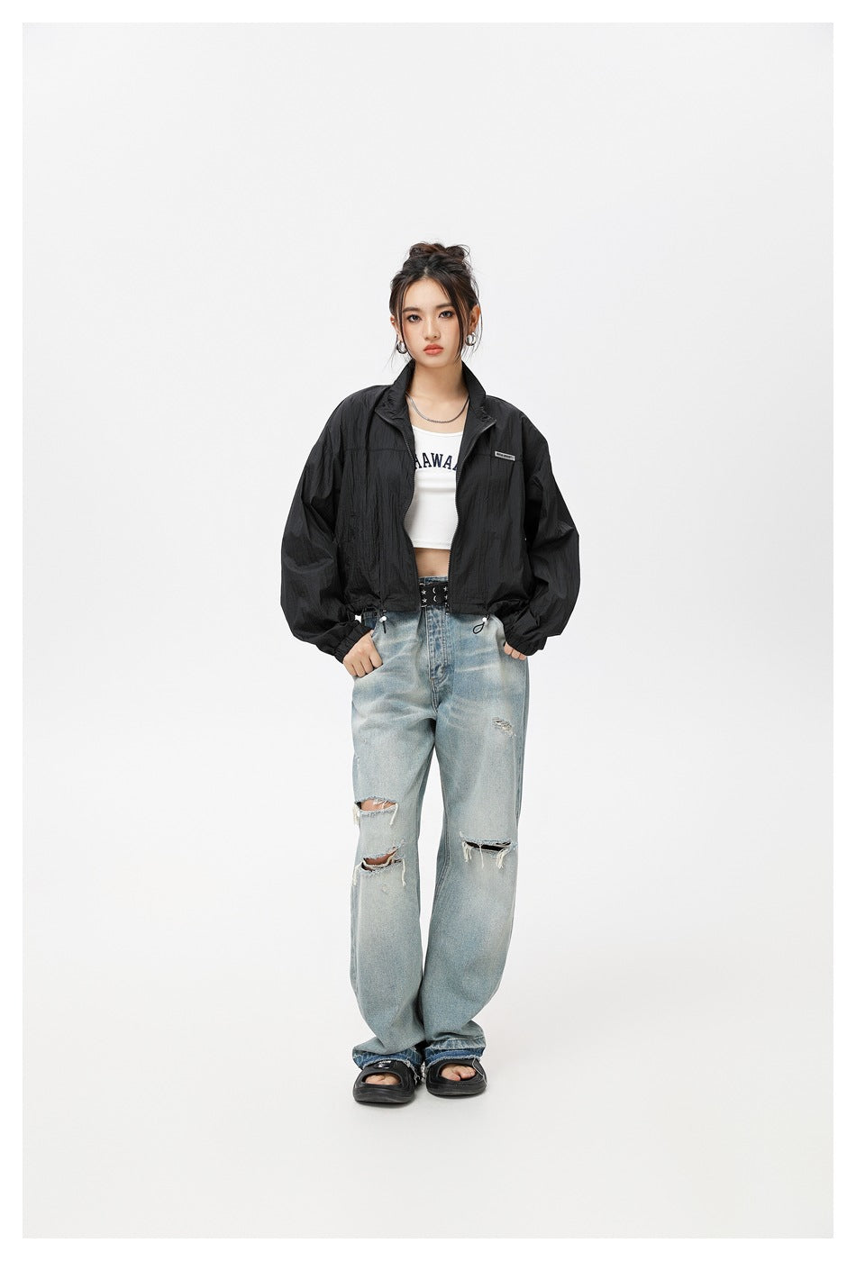 Model wearing a black cropped zip-up jacket over a white crop top with ripped light-wash jeans, full-length front view