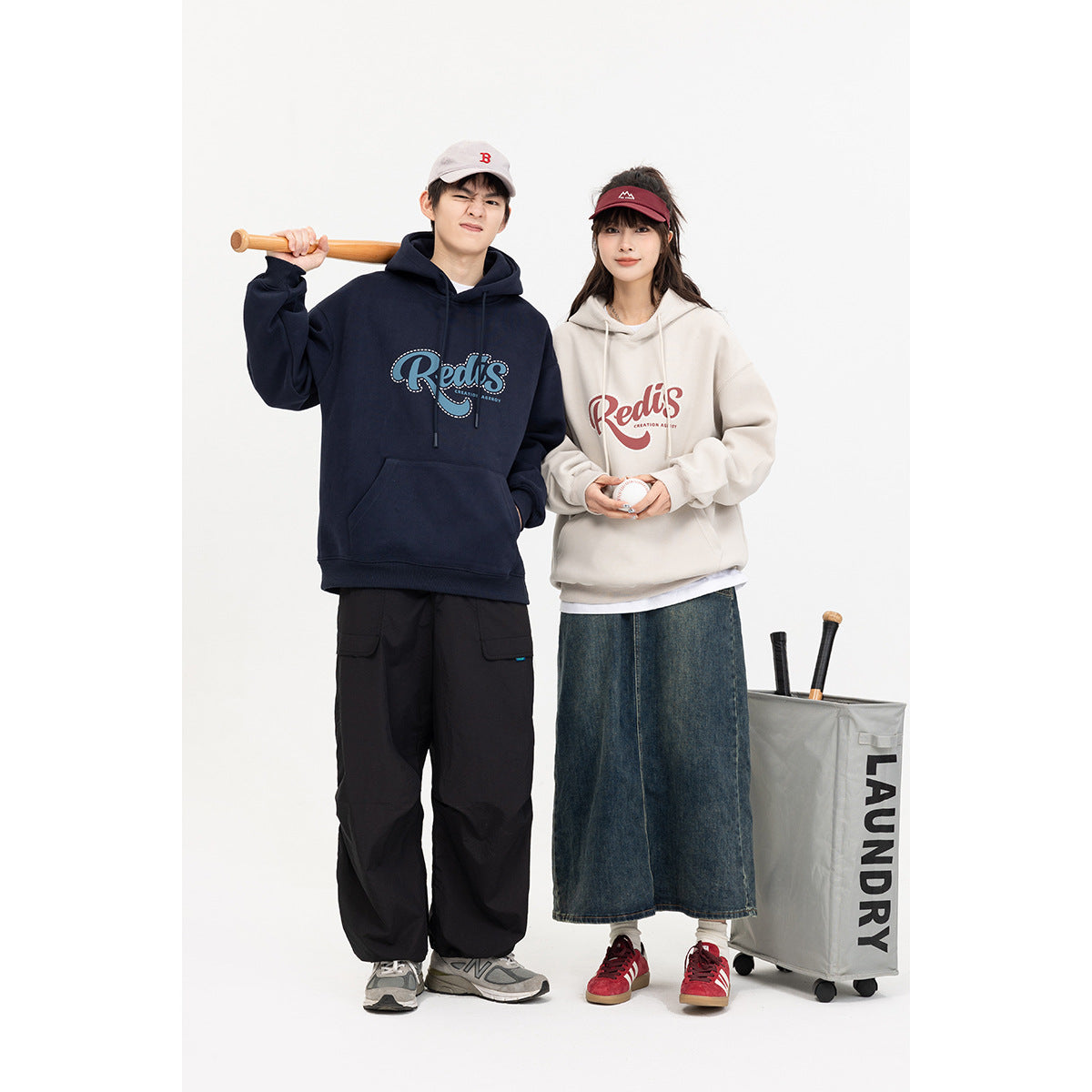 Two models wearing navy and cream graphic hoodies, posing with baseball bats and a ball.
