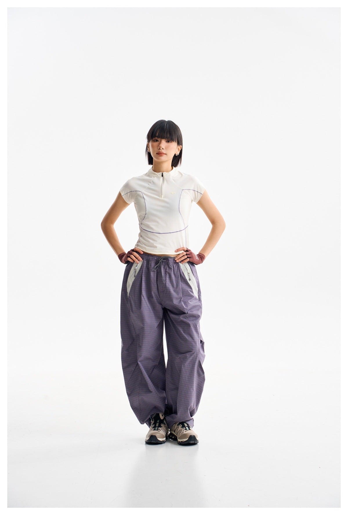 Model wearing an ivory cap-sleeve quarter-zip top with purple piping and loose purple plaid trousers.