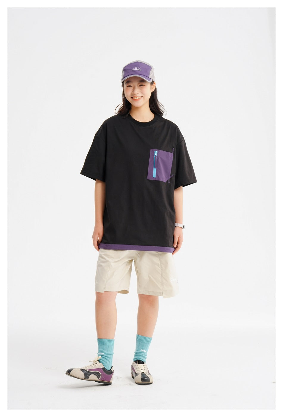 Person wearing a black T-shirt with a purple zippered chest pocket and purple hem, styled with beige shorts and a cap