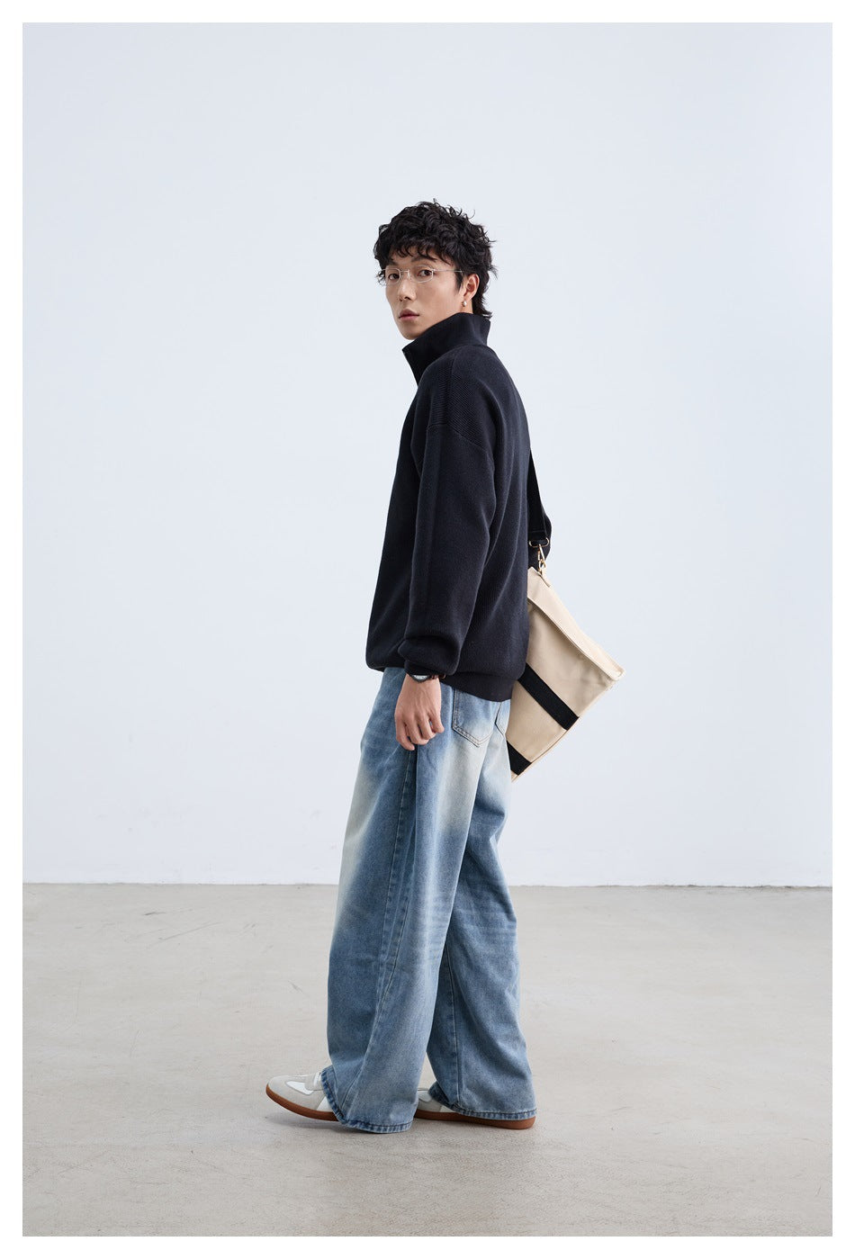 Side view of a model wearing a black quarter-zip sweater and loose blue jeans, with a beige tote over his shoulder.