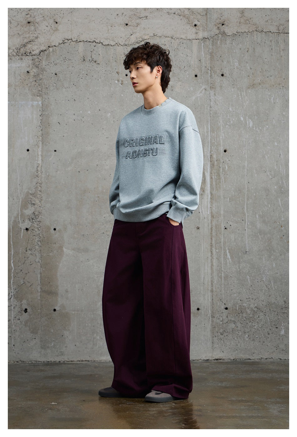 Model wearing a light gray crewneck sweatshirt with raised lettering and loose burgundy trousers, shown at an angle.